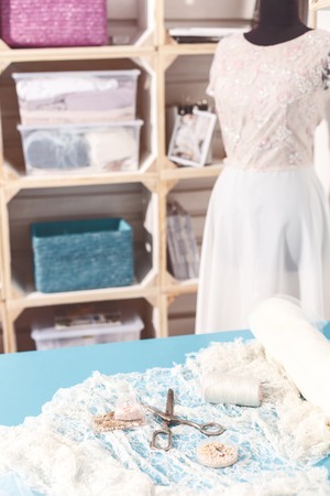 Close up of scissors, thread and beads on white cloth on the table. Shelves with material and wedding dress on background in atelierの写真素材