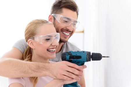 Drilling through the drywall. Smiling man and woman together making hole in white wall, using drill in special protective glassesの写真素材