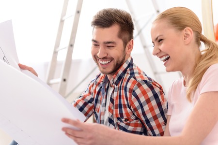 Very funny and amusing. Close up of attractive young couple is moving, holding paper with plan, discussing and smiling while looking sheetsの写真素材