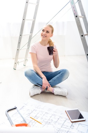 My day starts after coffee. Smiling girl sitting on floor with cup of coffee near her new apartment plan on background of ladderの写真素材