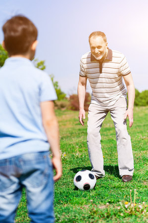 Cheerful grandfather and his grandchild are playing football in park. The man is preparing to kick the ball and smiling. The boy is standing and waitingの写真素材
