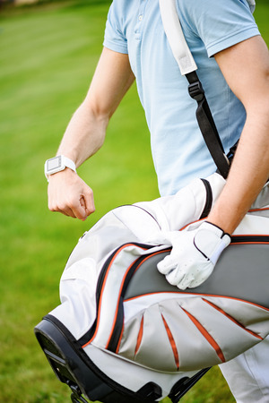 Exactly on time. Close up of young golfer checking time on beautiful golf course, holding his bag while walking to next holeの写真素材