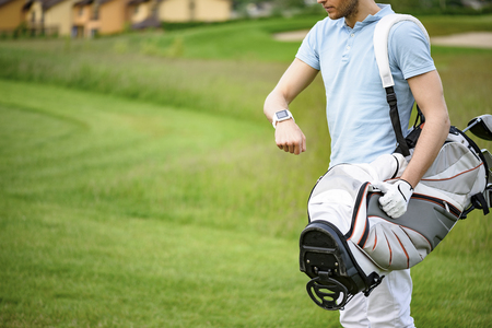 Very important not be late. Close up of golf player checking time on smartwatch while walking on beautiful golf course, carrying his bagの写真素材
