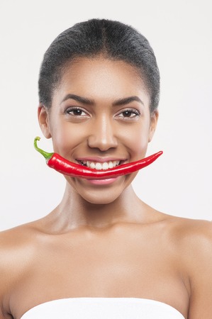 Portrait of pretty healthy african girl eating chili pepper. She standing and smiling. Isolatedの写真素材