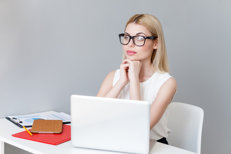Pretty young woman is making serious decision during work. She is sitting at desk near a laptop. The lady is looking forward pensivelyの写真素材