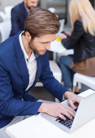 Cheerful young businessman is typing on laptop with inspiration. He is sitting at table in cafe and looking at screen seriously. Man and woman are communicating on backgroundの写真素材