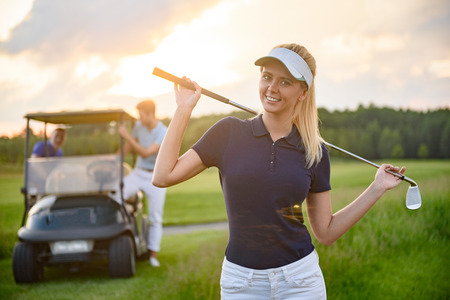 She enjoying golfing as hobby. Smiling young girl holding her golf club while on course with boyfriend and golf cart in backgroundの写真素材