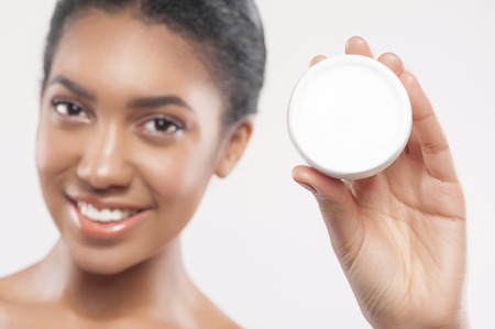 Beautiful mulatto girl is showing a can of cream to the camera. She is standing and smiling. Focus on the cosmetic product. Isolatedの写真素材