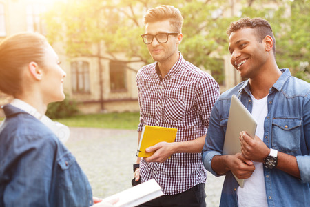 Happy young friends are talking and smiling. They are standing near university and holding learning...