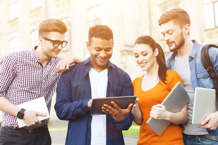Cheerful four students are enjoying university life. Young man is holding a tablet. His friends are...