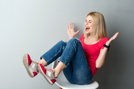 Attractive young woman is sitting on chair and laughing. She is gesturing with aspiration. Her eyes are closed with joy. Isolated on grey backgroundの写真素材