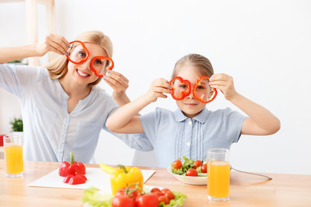 Having fun in kitchen. Smiling mother and daughter having fun, holding circles of red pepper near eyesの写真素材