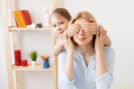 Family playtime. Little girl playfully putting her hands over eyes of her mother, playing at homeの写真素材