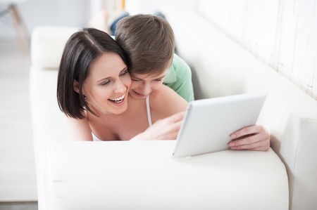 Motherly love. Top view of mother showing something on tablet to her adult son, lying on couchの写真素材