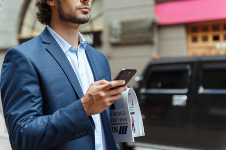 Confident young businessman is messaging on phone. He is standing and carrying newspaper outdoorsの写真素材