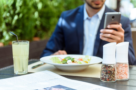 Young man is having lunch in restaurant. He is messaging on a mobile phone. Focus on plate of salad and drink on tableの写真素材