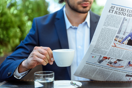 Successful young businessman is reading a newspaper and relaxing in restaurant. He is drinking coffee. Focus on cup and paperの写真素材