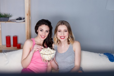 Cheerful friends are watching tv at home together. They are sitting on sofa and eating popcorn.の写真素材