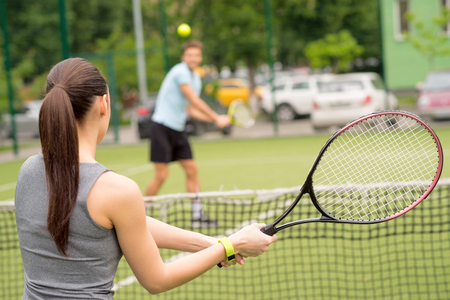 Cheerful woman and man are playing tennis on court. They are standing opposite the net and holding rackets. Focus on female backの写真素材