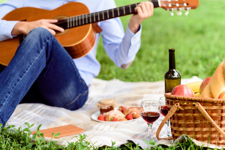 Romantic young man is playing guitar. He is sitting on blanket near food and drink in natureの写真素材