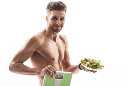 Eat vegetables and lose weight. Cheerful young man is showing scales and plate of salad. He is looking at camera and smiling. Isolatedの写真素材