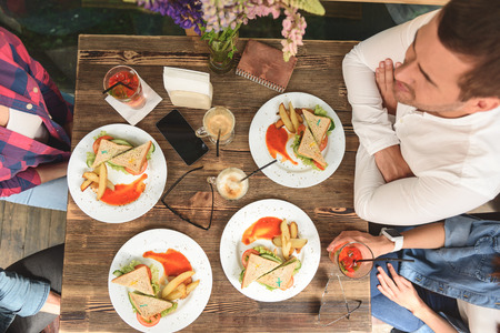 Food brings friends together. Top view of wooden table with plates of sandwiches, latte, cocktails, glasses, mobile phone, notebookの写真素材