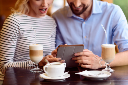 Cheerful friends are having fun in cafe. They are looking at tablet with interest and smiling. Man and woman are sitting at desk near cups of latteの写真素材