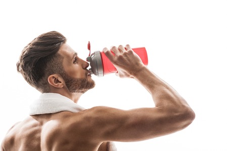 Cheerful young man is drinking water from sporty bottle. He is standing with relaxation. Athlete is holding towel over neck. Isolatedの写真素材