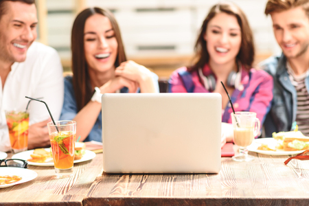 Sharing some time online. Close up of laptop standing on wooden table with cheerful four smiling friends in backgroundの写真素材