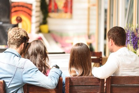 All together again. Back view of four friends sitting on wooden chairs in cafeteria and watching somewhereの写真素材