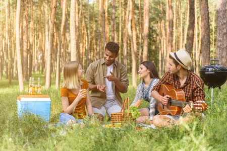 Happy men and women are talking and laughing in the nature. They are sitting on grass. Man is playing guitar with enjoymentの写真素材