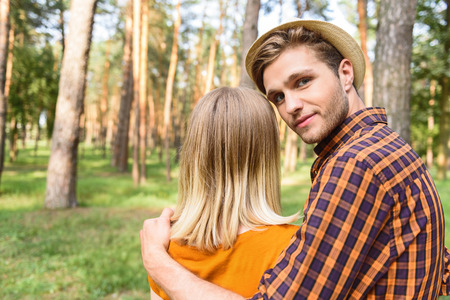 Handsome young man is embracing woman with support. He is looking at camera with confidence. Lovers are standing in forestの写真素材