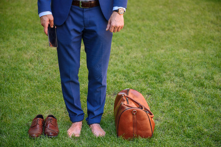 Close up of male legs standing on grass barefoot. Man is holding mobile phone. There are his bag and shoes on lawnの写真素材
