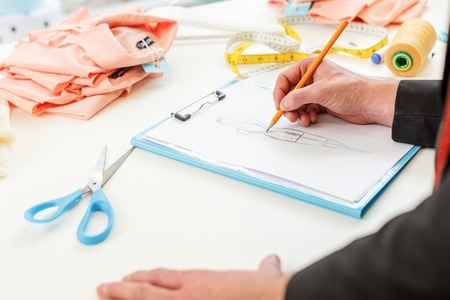 Close up of male hand drawing sketch of dress. Designer is standing and leaning on table in atelierの写真素材