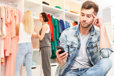Young man is bored while his girlfriend is choosing clothing in shop. He is sitting and using mobile phoneの写真素材
