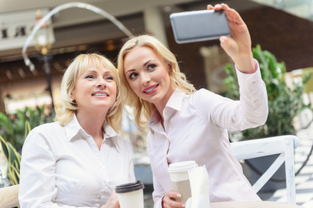 Happy woman is making selfie with her parent. She is holding mobile phone and cup of coffee. Ladies are sitting at table in cafeの写真素材