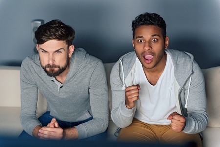 Tense moment. Two young handsome men looking at something dramatic on television, sitting on sofa, isolated on grey backgroundの写真素材
