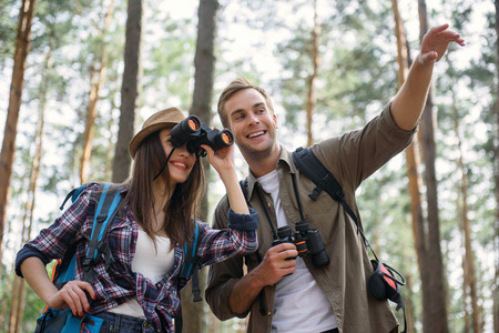 Happy loving couple is making journey in forest. Man is showing something and smiling. Woman is looking into binoculars with interestの写真素材