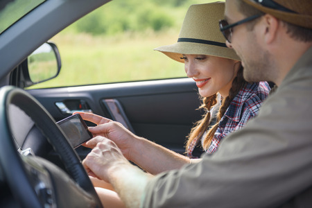 Happy loving couple is traveling by car on vacation. Man is showing mobile phone to his girlfriend. Woman is looking at map and smilingの写真素材