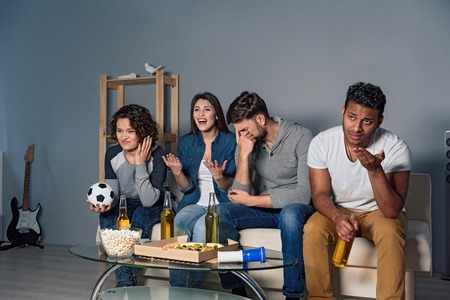 We do not believe in defeat. Friends upset watching football match, sitting on white couch near gray wallの写真素材