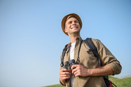 Joyful male tourist is hiking on meadow. He is standing and smiling. Man is holding binoculars