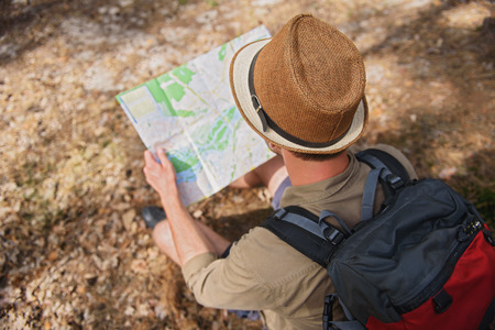 Young male tourist is looking for place of destination. He is sitting on ground in forest and...