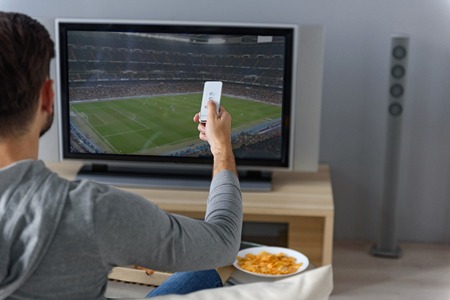Choosing favorite channel. Close up of man sitting back and holding remote, switching channel on football gameの写真素材