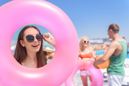 Pretty girl is looking through rubber ring with interest. She is adjusting sunglasses and laughing. Her friends are standing near swimming pool and talkingの写真素材