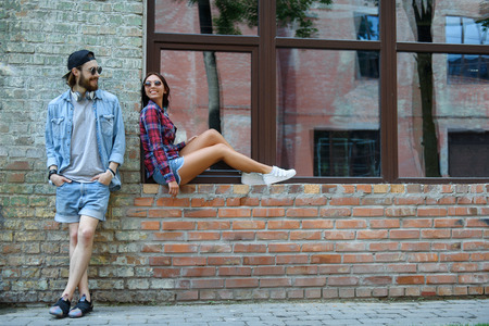 Carefree couple is talking and smiling. Woman is sitting on windowsill of building. Man is standing and looking at her flirtinglyの写真素材