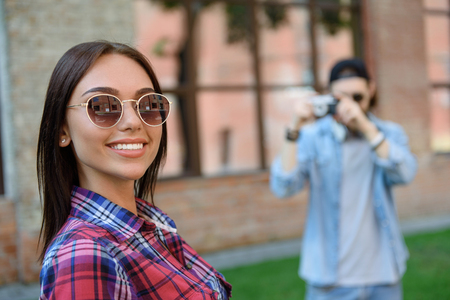 Happy young woman is looking at camera and smiling. Her boyfriend is standing and photographing her on backgroundの写真素材