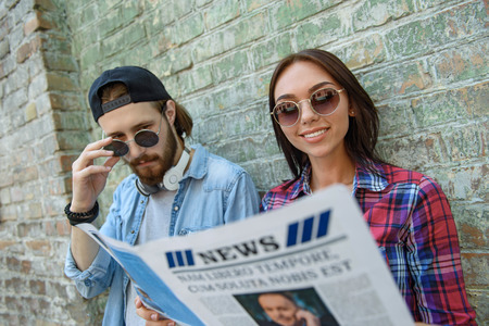 Joyful friends are reading paper outdoors. Woman is standing and smiling. Man is looking at article with interestの写真素材