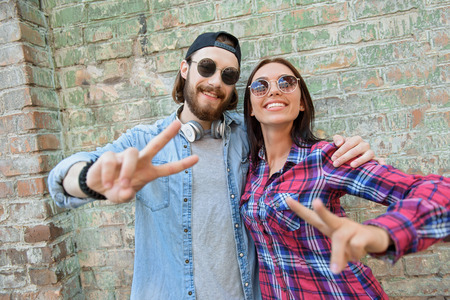 Pretty couple is showing peace sign and smiling. They are standing and embracing. Man and woman are wearing sunglassesの写真素材