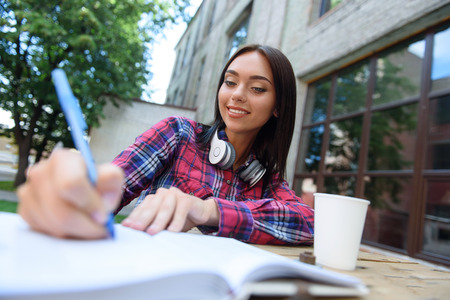 Happy young student is studying in campus. She is writing into notebook and smiling. Girl is sitting at desk outdoorsの写真素材
