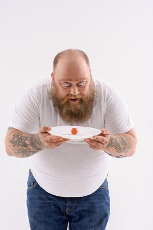 Only one tomato. Thick man is dieting. He is holding plate and looking at vegetable with surprise. Isolatedの写真素材